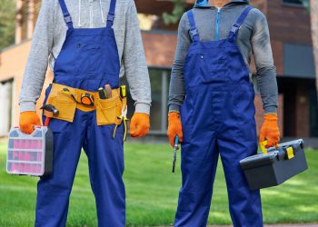 Cropped shot of two male builders in blue overalls carrying toolbox at construction site. Building, protective gear and people concept