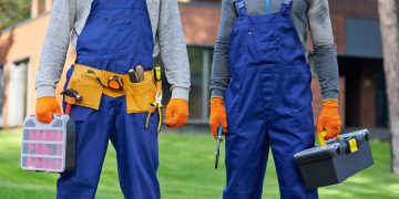 Cropped shot of two male builders in blue overalls carrying toolbox at construction site. Building, protective gear and people concept