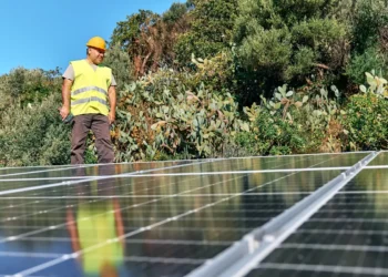 A man with short brown hair in a yellow hardhat and vest stands near solar panels on a sunny day.